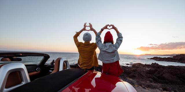 Couple enjoying beautiful views on the ocean, standing together near the car on the rocky coast, showing with hands heart shape. Carefree lifestyle, love and travel concept