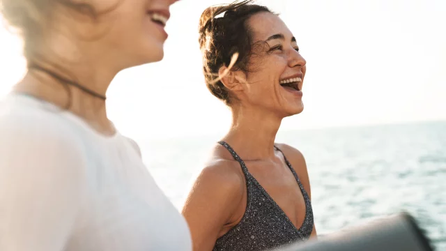 Image of happy fitness women in sportive clothes smiling and standing with yoga mat by seaside in morning