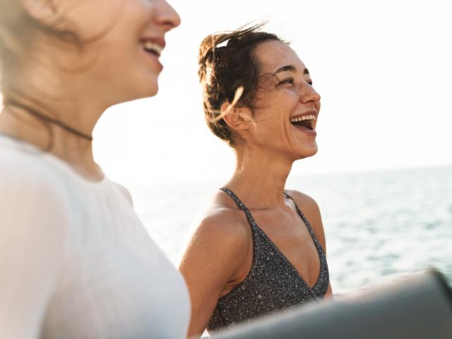 Image of happy fitness women in sportive clothes smiling and standing with yoga mat by seaside in morning